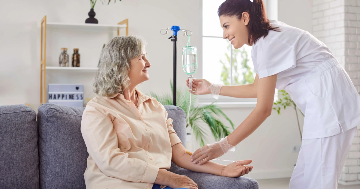 An older woman with grey hair smiles as a nurse in white prepares an IV drip in a bright, home-like setting for IV Therapy in St. George, UT.