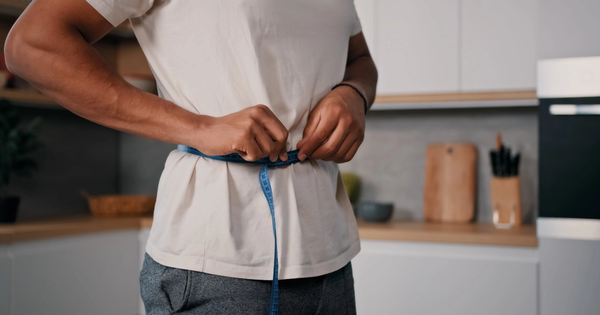 A person in a kitchen measuring their waist with a blue measuring tape over a white t-shirt, representing Medical Weight Loss in St. George, UT.