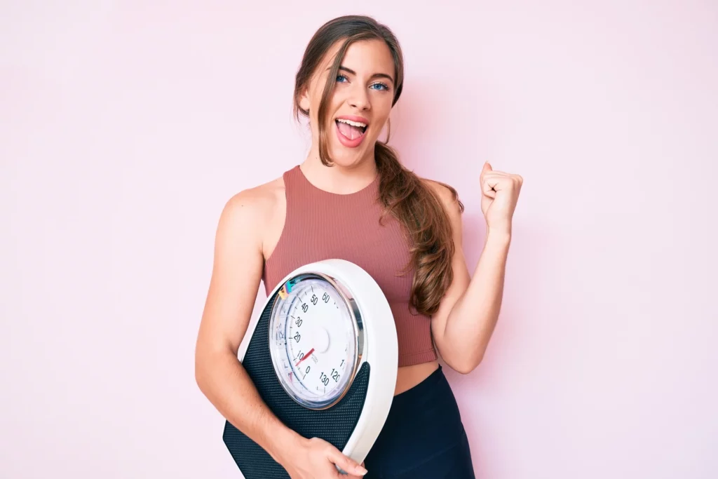Woman happy after achieving weight loss goals and holding body weighing machine | Mesa Medical Health & Wellness | St. George, UT
